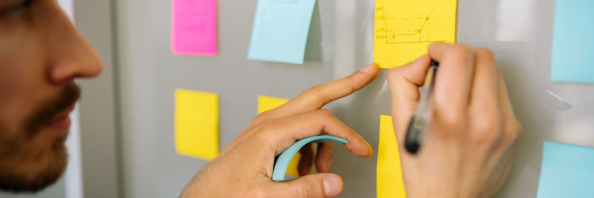 A close-up of a man's face as he draws a growth chart on a yellow sticky note attached to a glass wall.